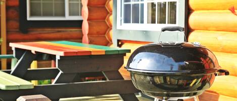 A barbecue grill in front of a wooden cabin with a window and a picnic table.