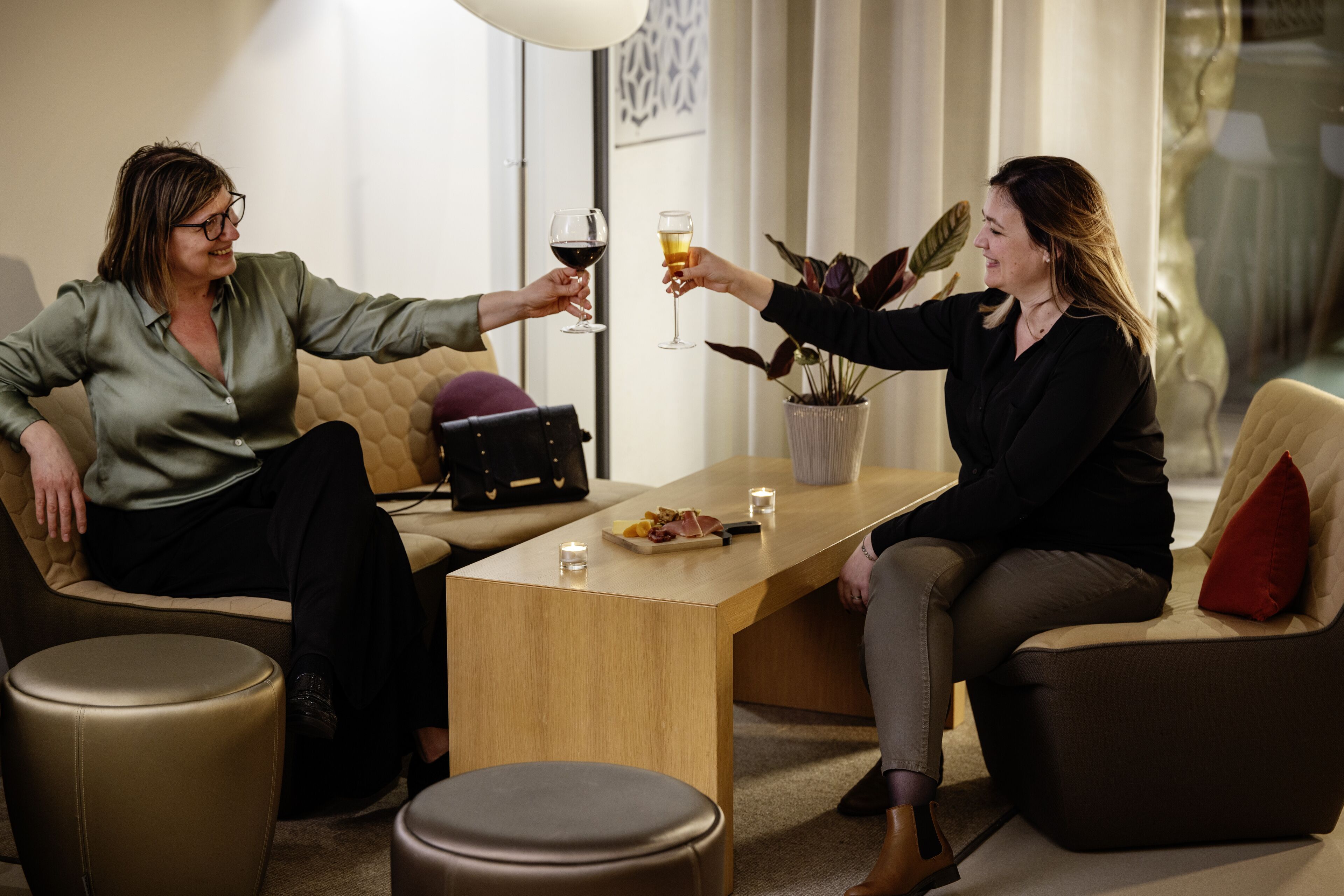 Deux personnes sont assises sur un canapé et une chaise, et portent un toast avec des verres à vin. Une petite table, sur laquelle se trouvent une assiette de nourriture et une bougie, est placée entre elles.