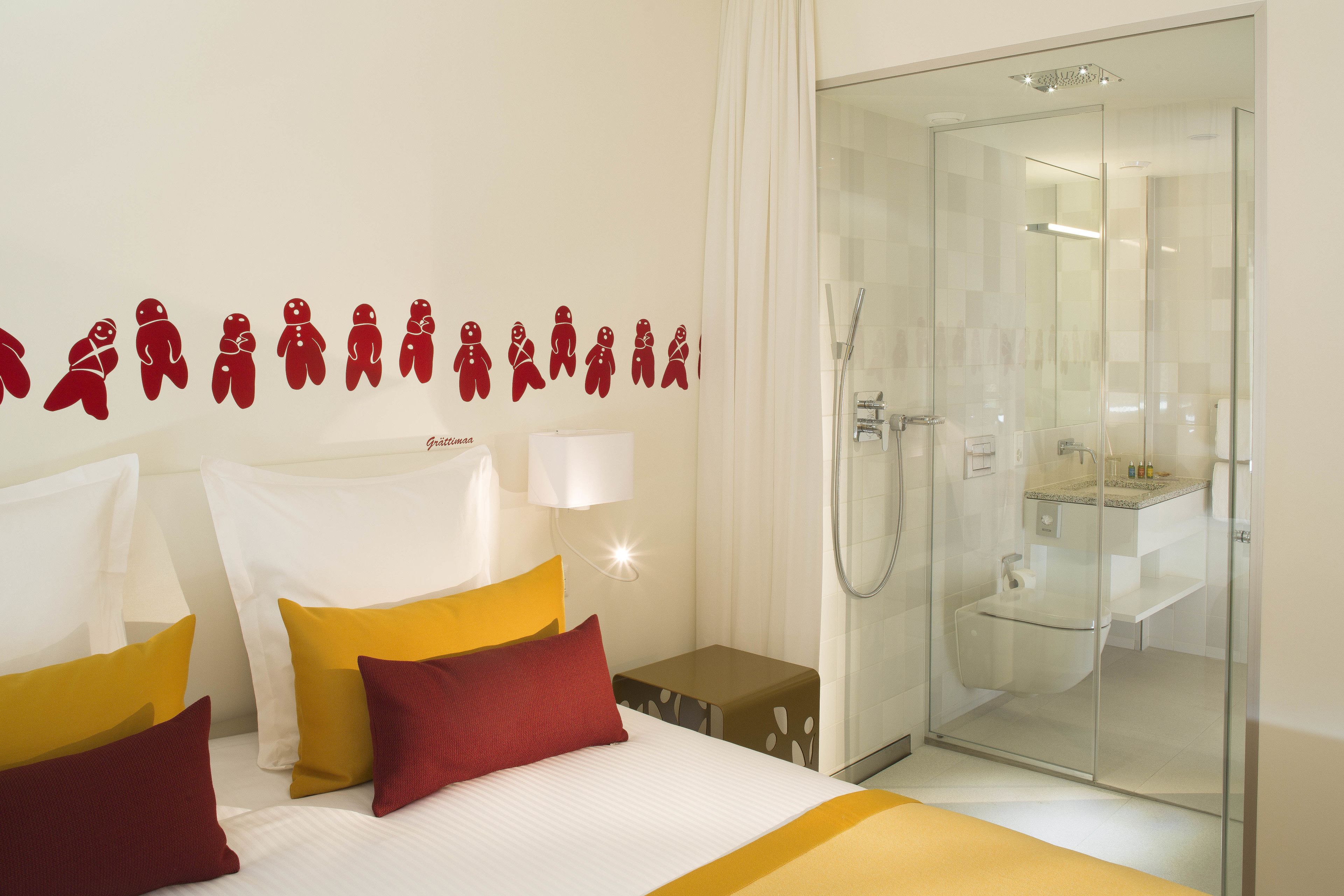 A hotel room with a bed, pillows, a nightstand, a wall with gingerbread man decals, a glass shower enclosure, and a bathroom with a toilet and sink.