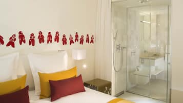 A hotel room with a bed, pillows, a nightstand, a wall with gingerbread man decals, a glass shower enclosure, and a bathroom with a toilet and sink.