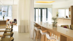 A modern restaurant with a long wooden bar, white chairs, and a large window with curtains.
