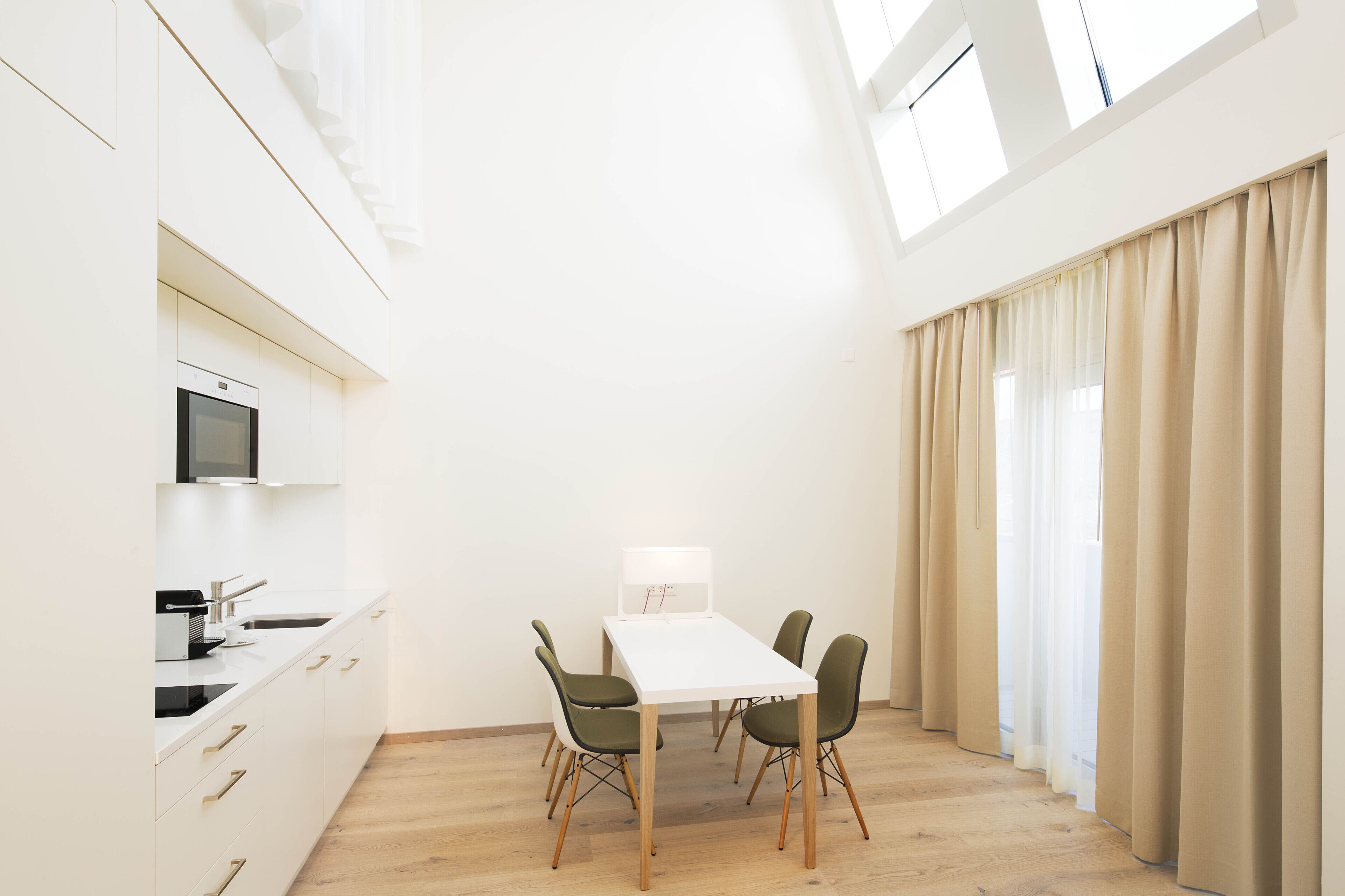 A modern kitchen with white cabinets, a microwave, and a sink. There is a dining table with four chairs and a lamp. The room has wooden flooring and large windows with beige curtains.