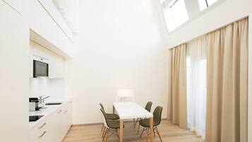 A modern kitchen with white cabinets, a microwave, and a sink. There is a dining table with four chairs and a lamp. The room has wooden flooring and large windows with beige curtains.