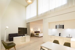 A modern, well-lit room with a flat-screen TV, a sofa, a dining table, and a kitchenette.