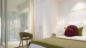 A modern hotel room with a glass shower enclosure, a single bed with green bedding, and a green chair.