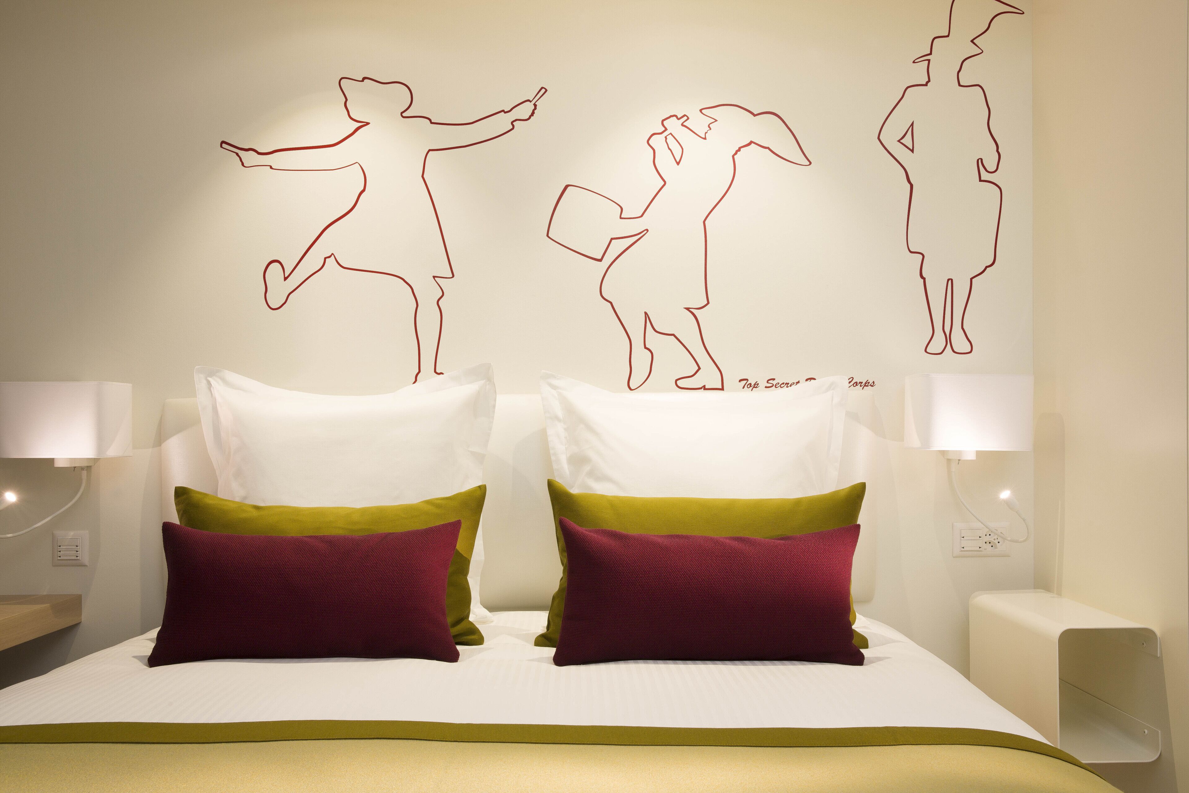 A neatly made bed with white and green pillows, a wall with red line art of people, and a white nightstand with a lamp.