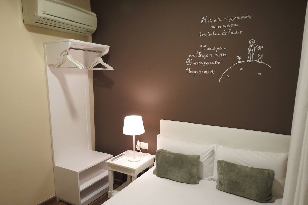 A hotel room with a bed, a bedside table with a lamp, a hanging coat rack, and a wall with motivational text.