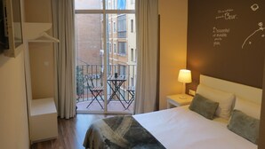 Une chambre d’hôtel avec un lit, une petite table avec une lampe, un balcon avec des chaises et une vue sur les bâtiments environnants.