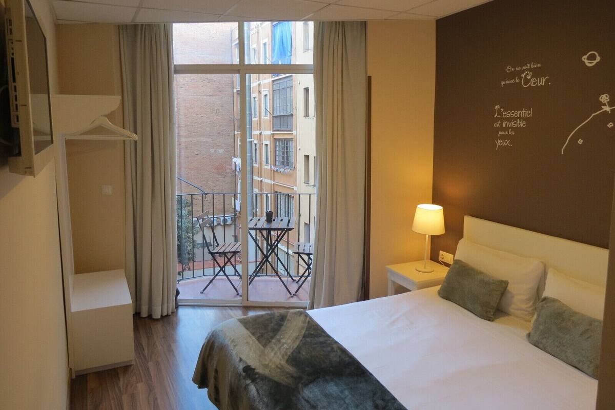A hotel room with a bed, a small table with a lamp, a balcony with chairs, and a view of buildings outside.