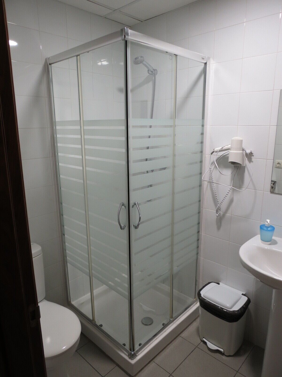 Ducha con cerramiento de vidrio, cabezal de ducha de mano, inodoro blanco y lavamanos con dispensador de jabón.