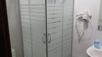 Ducha con cerramiento de vidrio, cabezal de ducha de mano, inodoro blanco y lavamanos con dispensador de jabón.