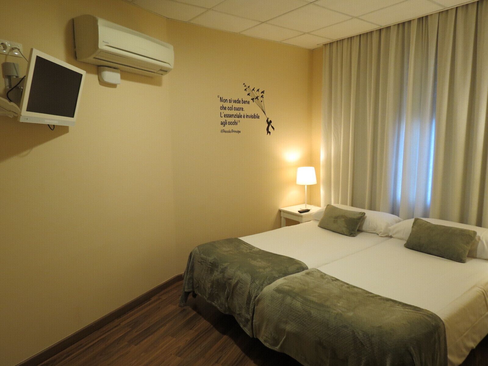 A hotel room with a bed, a desk with a lamp, an air conditioner, and a wall decoration.