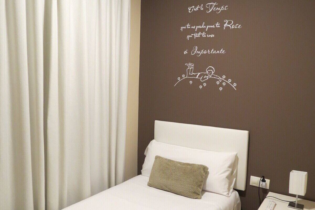 A hotel room with a bed, a nightstand, a lamp, and a wall with decorative text.