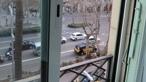Une vue depuis une fenêtre ornée d’une rambarde en métal, donnant sur une rue animée par la circulation des voitures et bordée d’immeubles.