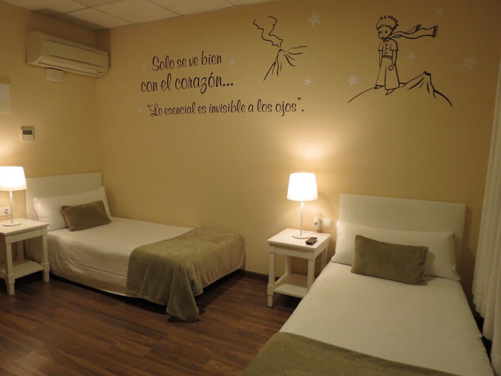 A hotel room with two beds, bedside tables, lamps, and a wall with a quote and illustrations.