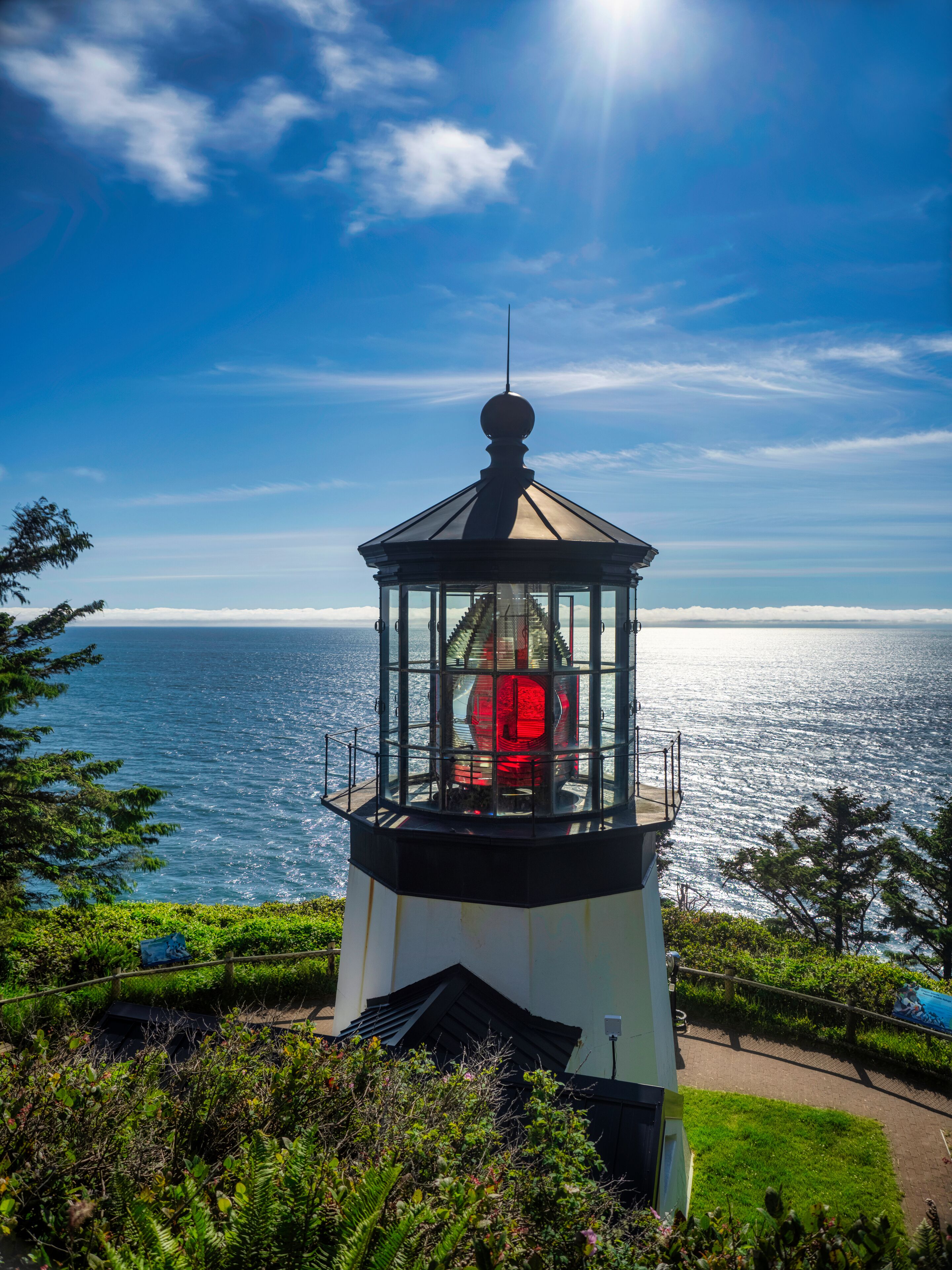 Un faro con cerramiento de vidrio, rodeado de vegetación y con vistas al mar.