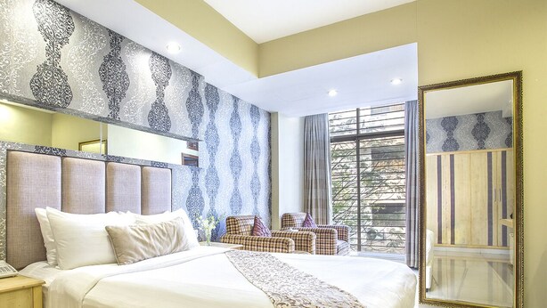 Camera d'albergo con un letto grande, una testiera dal motivo decorativo, un angolo salotto con divano e poltrone, uno specchio ampio e una finestra con tende.