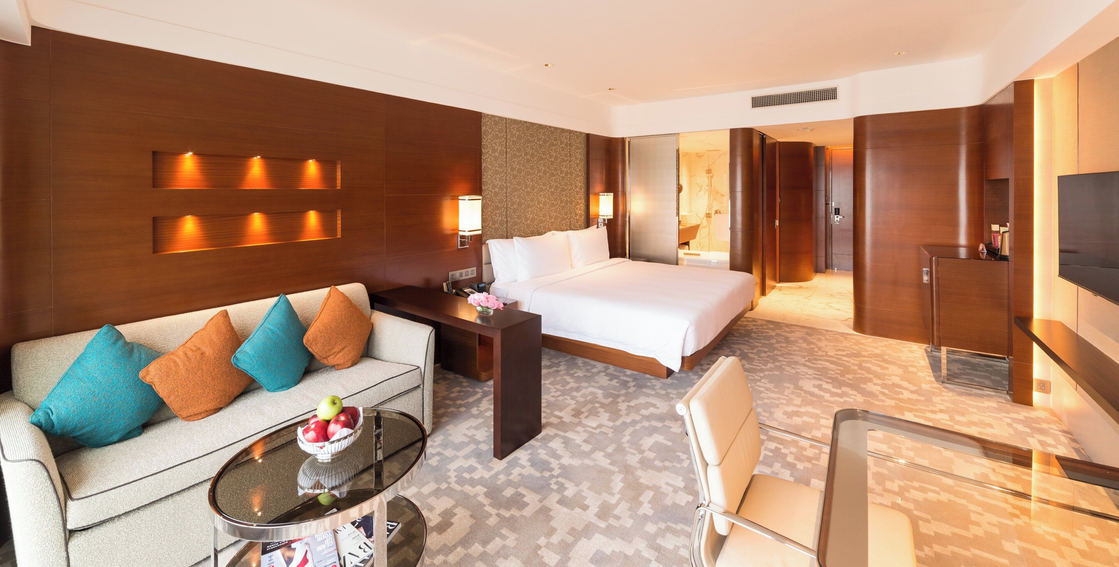 A hotel room with a bed, a sofa, a coffee table with a bowl of fruit, a desk, and a TV.