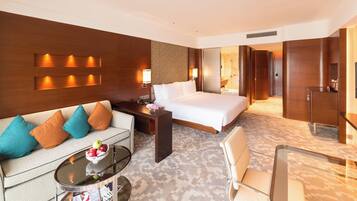 A hotel room with a bed, a sofa, a coffee table with a bowl of fruit, a desk, and a TV.