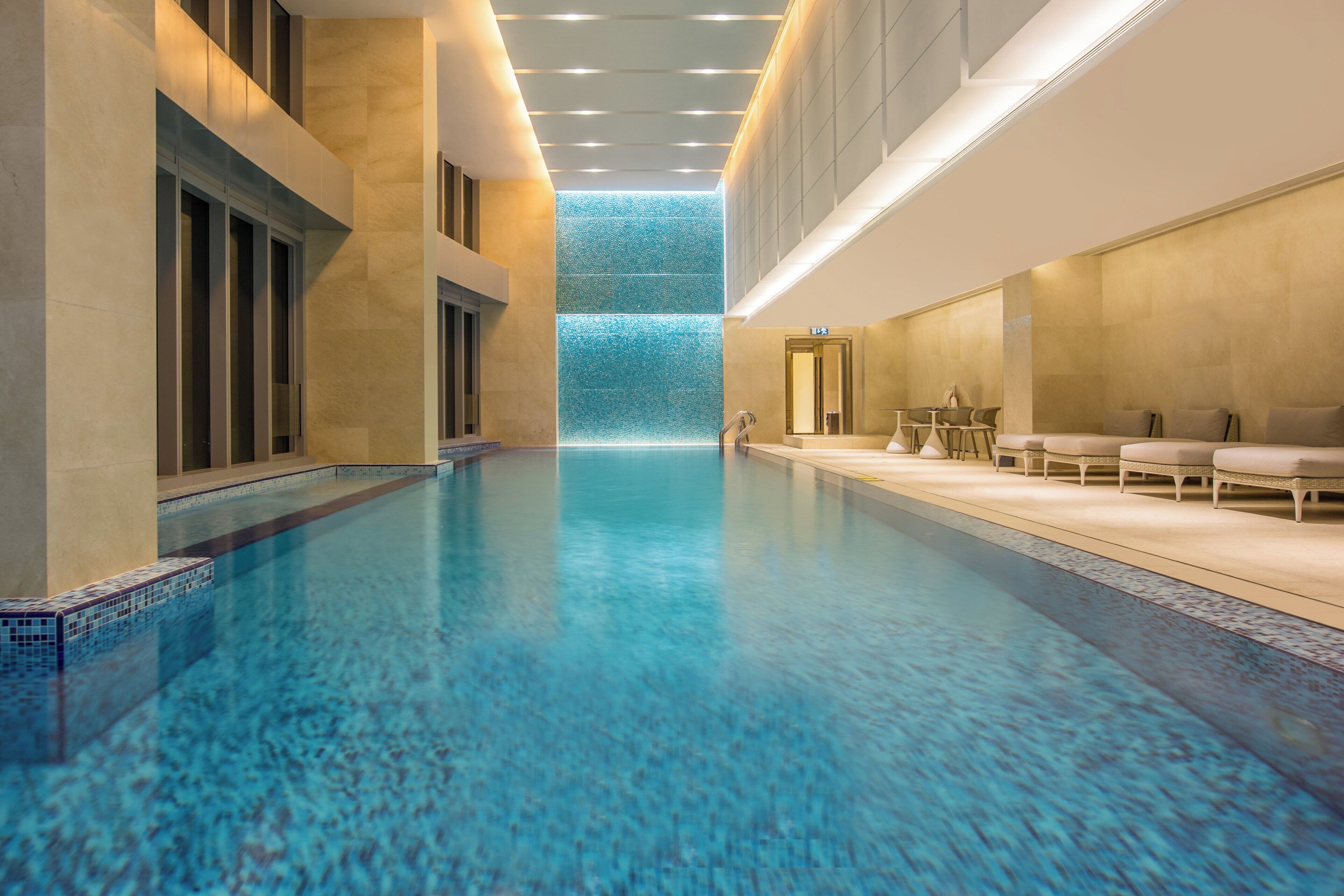 A hotel pool area with a clear view of the pool, a seating area with chairs and tables, and a glass wall with a view of the sky.