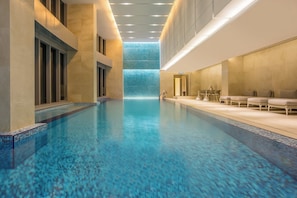 Un área de piscina de hotel con vista despejada, una zona de estar con sillas y mesas, y un muro de cristal con vista al cielo.