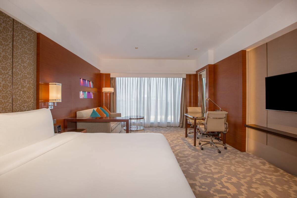 Ein Hotelzimmer mit einem großen Bett, einem Schreibtisch mit Stuhl, einem Fernseher und einem Fenster mit Vorhängen.