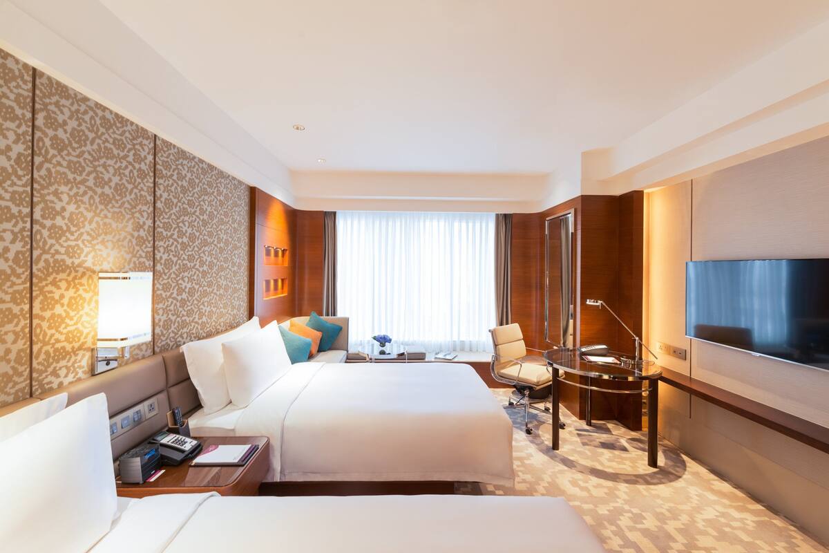 Ein Hotelzimmer mit einem großen Bett, einem Schreibtisch mit Lampe, einem Fernseher und einem Fenster mit Vorhängen.