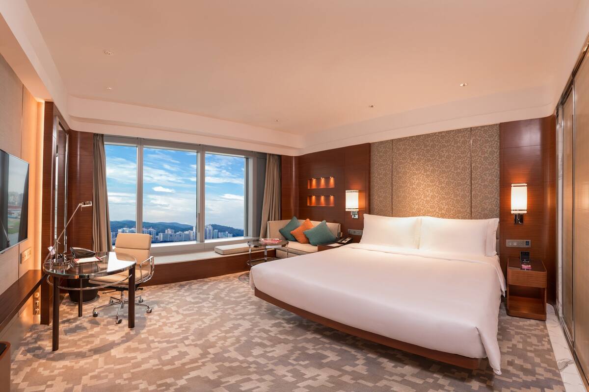 Geräumiges Hotelzimmer mit einem großen Bett, einem Schreibtisch und einem Stuhl. Ein großes Fenster bietet Blick auf die Stadt.