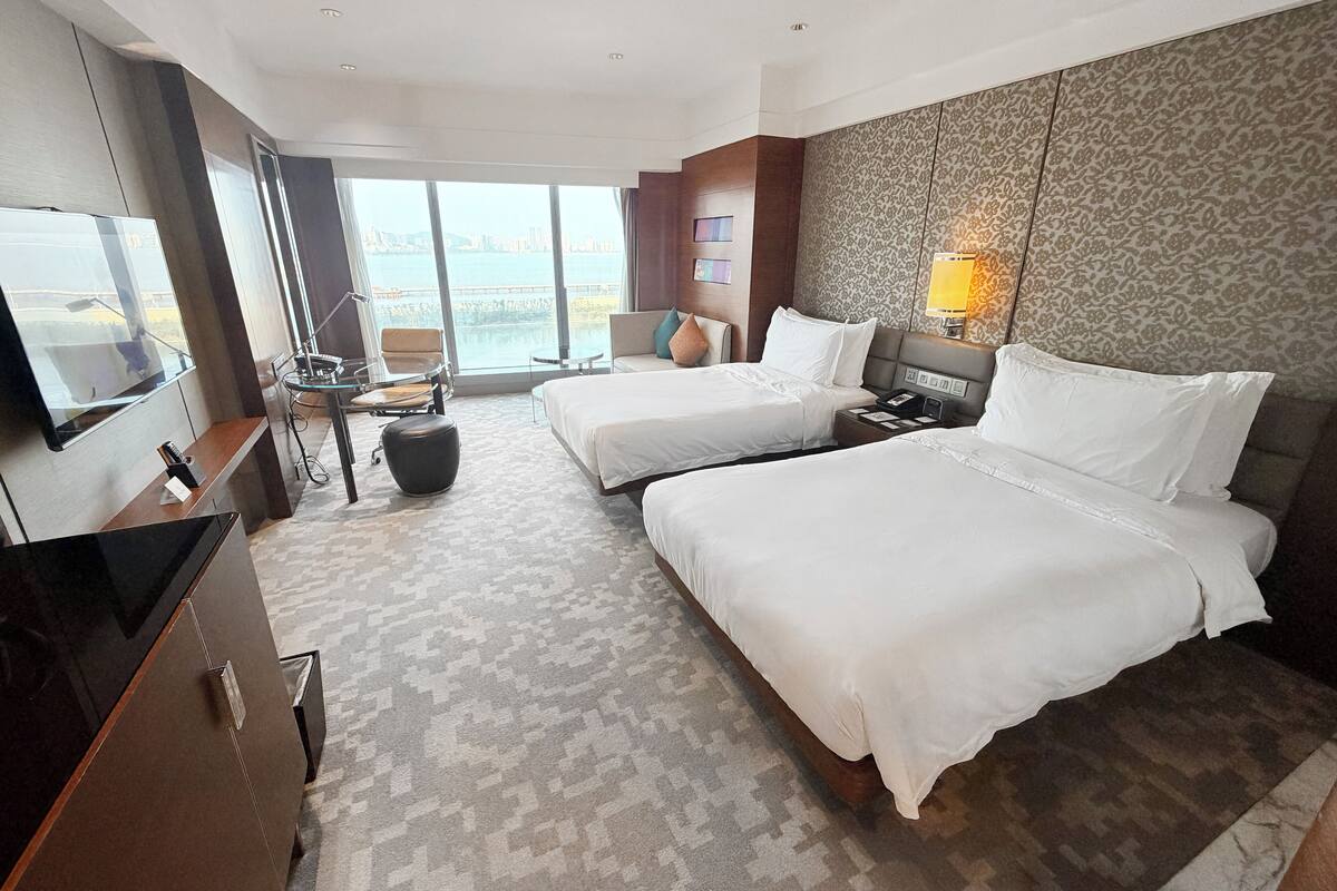 Ein Hotelzimmer mit zwei Betten, einem Fernseher, einem Schreibtisch, einem Stuhl und Meerblick.