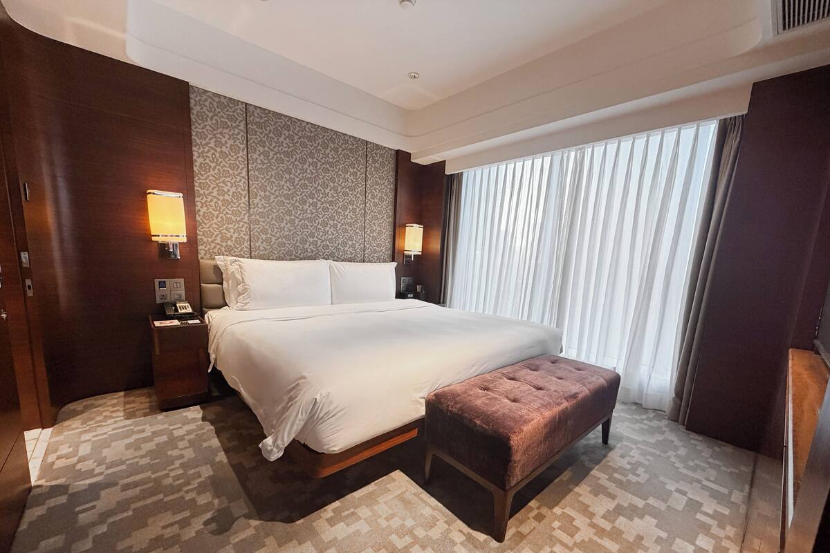 Ein Hotelzimmer mit einem großen Bett, einem Nachttisch mit Lampe, einem kleinen Schreibtisch und einem großen Fenster mit Vorhängen.