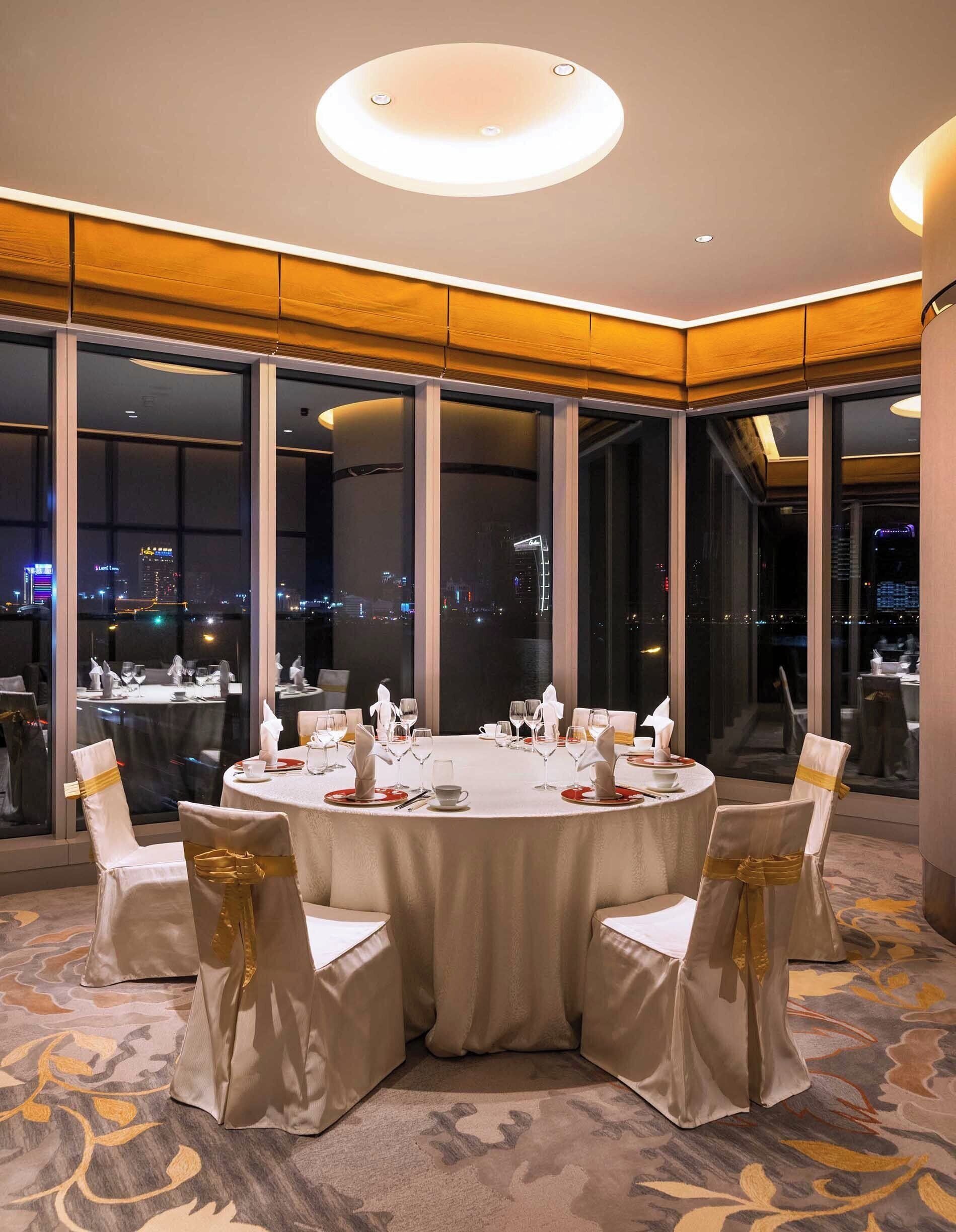 A round dining table with chairs, set for a meal, in a room with large windows offering a city view.
