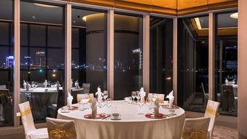A round dining table with chairs, set for a meal, in a room with large windows offering a city view.