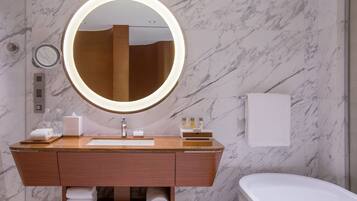 A bathroom with a marble wall, a wooden vanity, a round mirror with a light, a white sink, and a white bathtub.