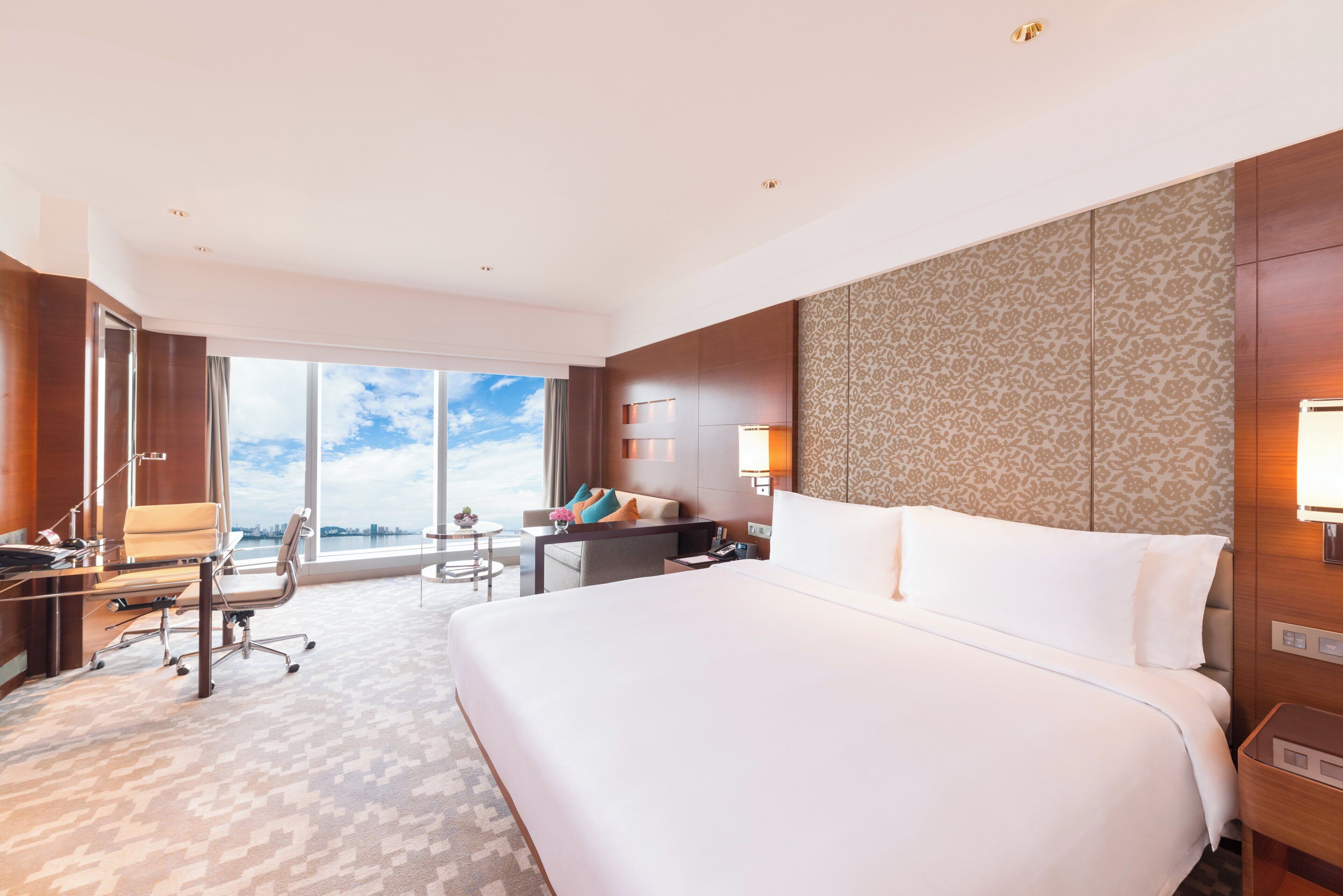 A hotel room with a large bed, a desk with a chair, a small table, and a view of the city through large windows.
