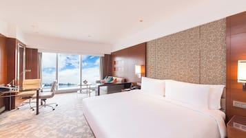 A hotel room with a large bed, a desk with a chair, a small table, and a view of the city through large windows.