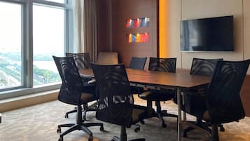 Een vergaderruimte met een grote tafel, zes stoelen, een televisie aan de muur en een stadsgezicht door grote ramen.