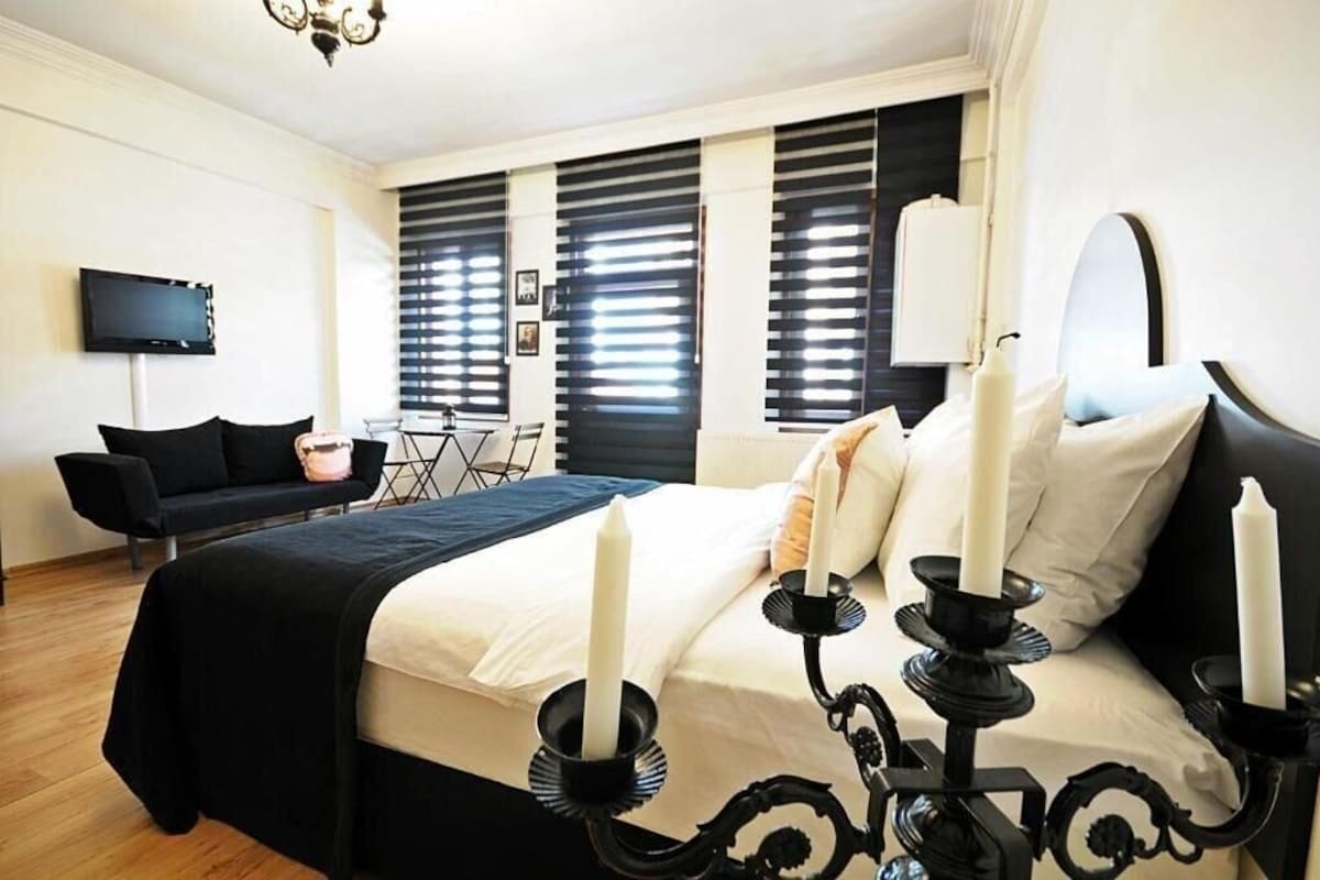 A bedroom with a bed, black and white bedding, a TV, a black sofa, and a chandelier.