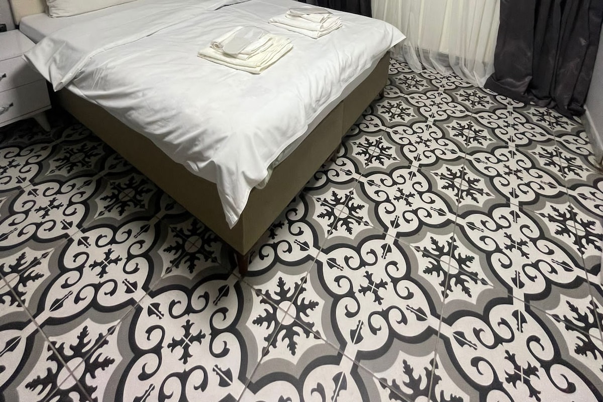 A neatly made bed with white linens, a bedside table with a lamp, and a patterned floor.