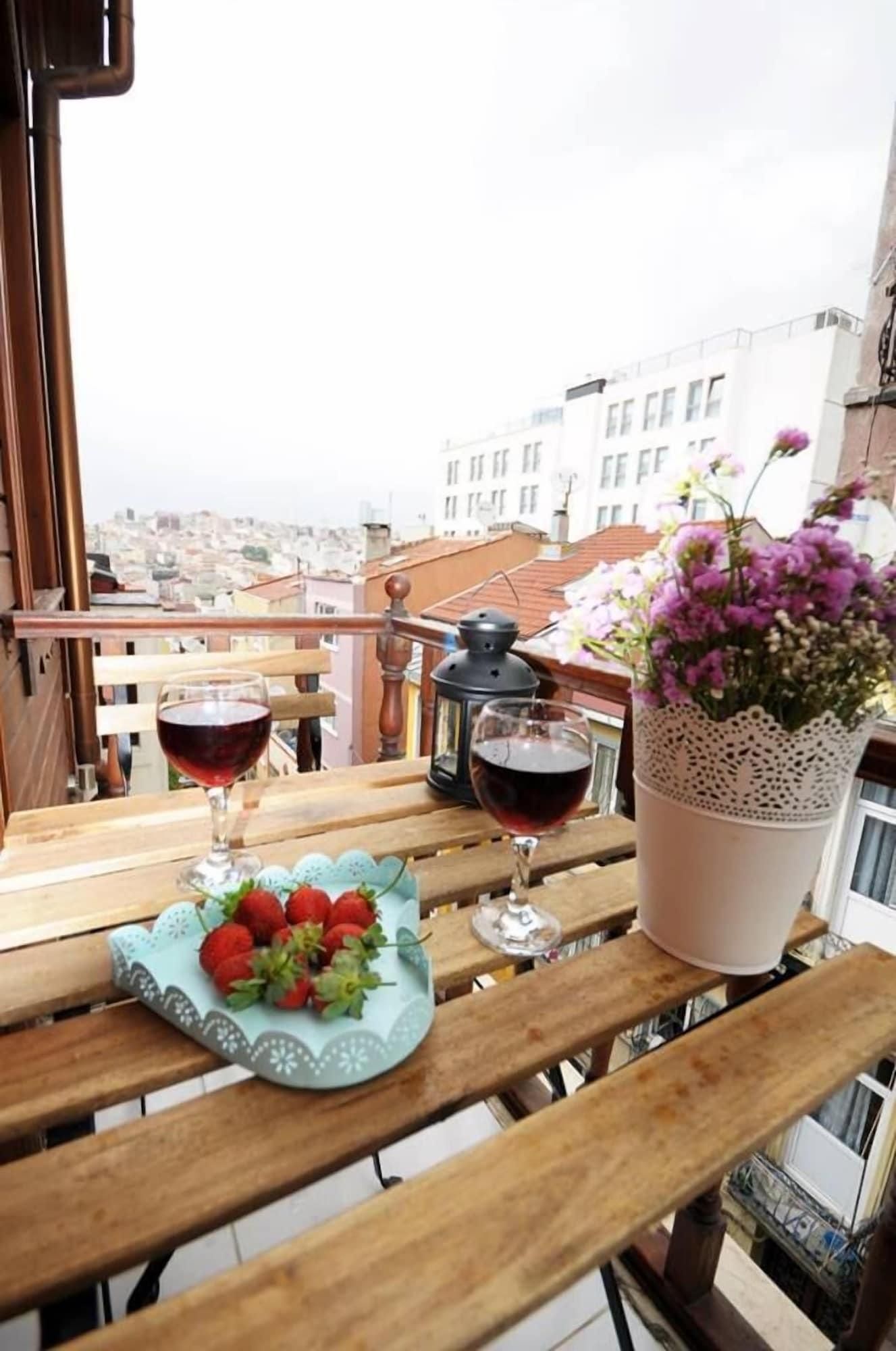 Una mesa de madera con dos copas de vino, un plato de fresas y una planta en maceta con flores moradas en un balcón con vistas a un paisaje urbano.