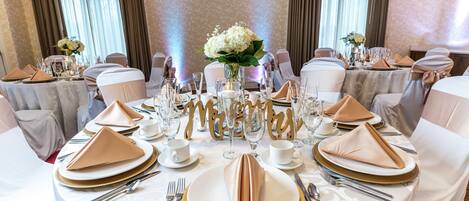 A formal dining room with round tables, white tablecloths, gold-rimmed plates, and white chairs.