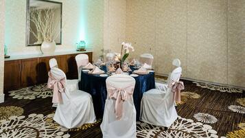 Een balzaal met ronde tafels gedekt voor een formeel evenement, met witte stoelen met roze linten, een centraal bloemstuk en een kamerbreed tapijt met patroon.