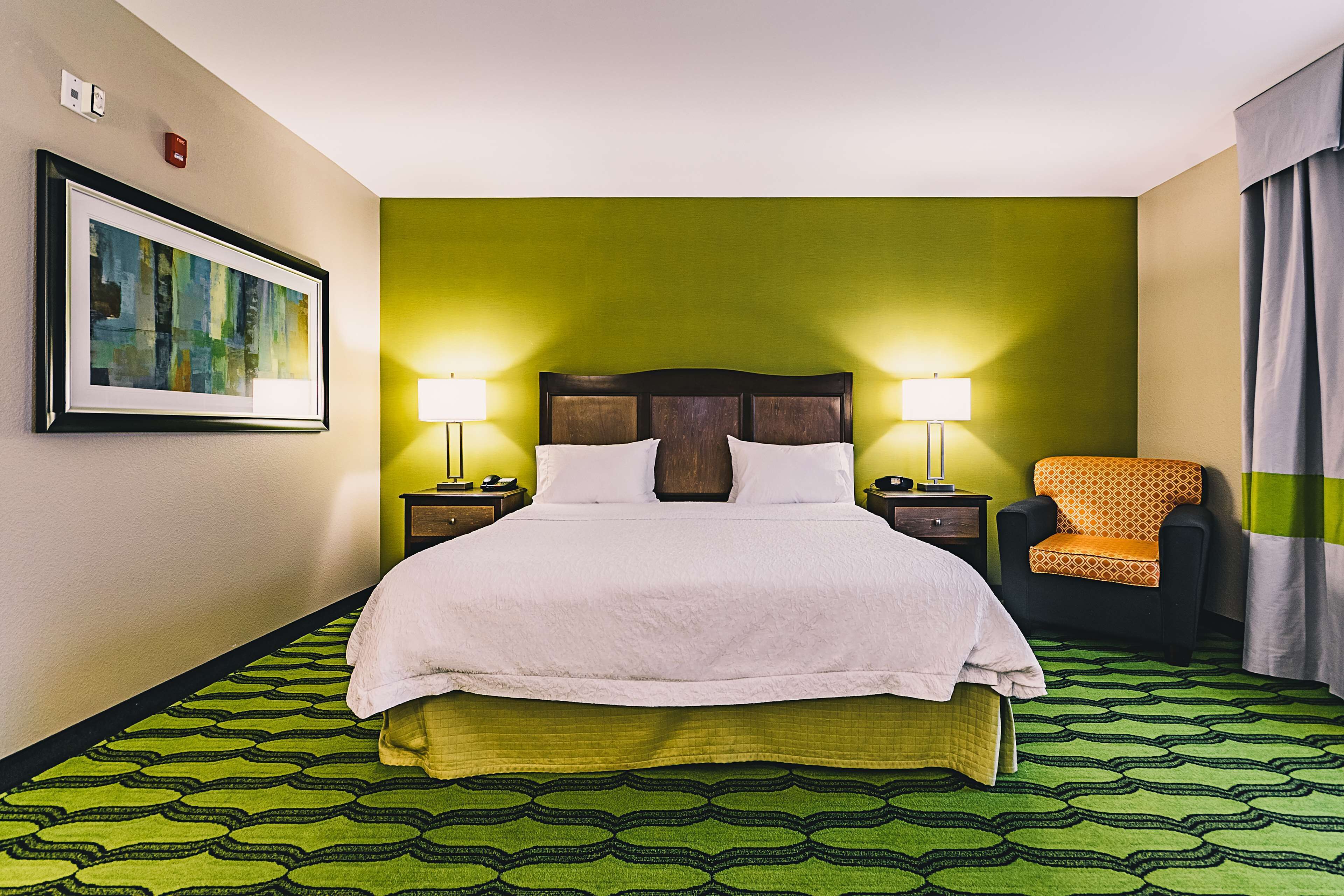 A hotel room with a bed, two bedside lamps, a chair, a framed picture on the wall, and a patterned carpet.