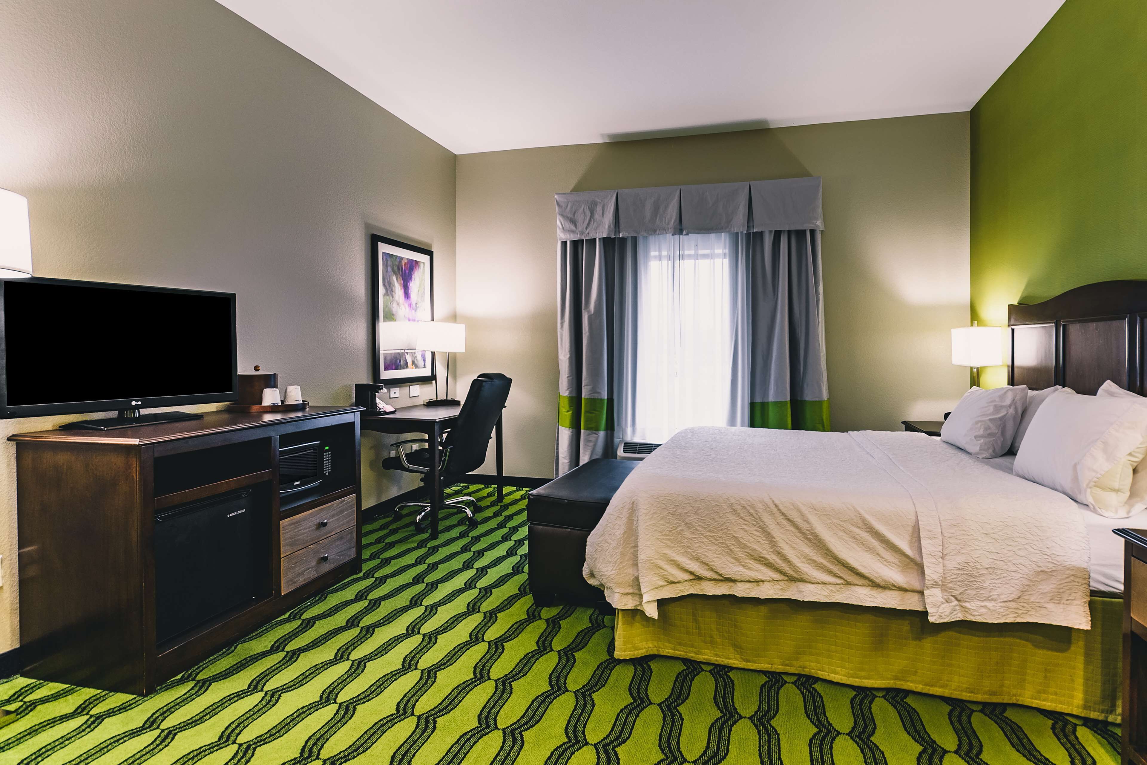 Ein Hotelzimmer mit einem Bett, einem Schreibtisch mit Computer, einem Fernseher und einem grün gemusterten Teppich.