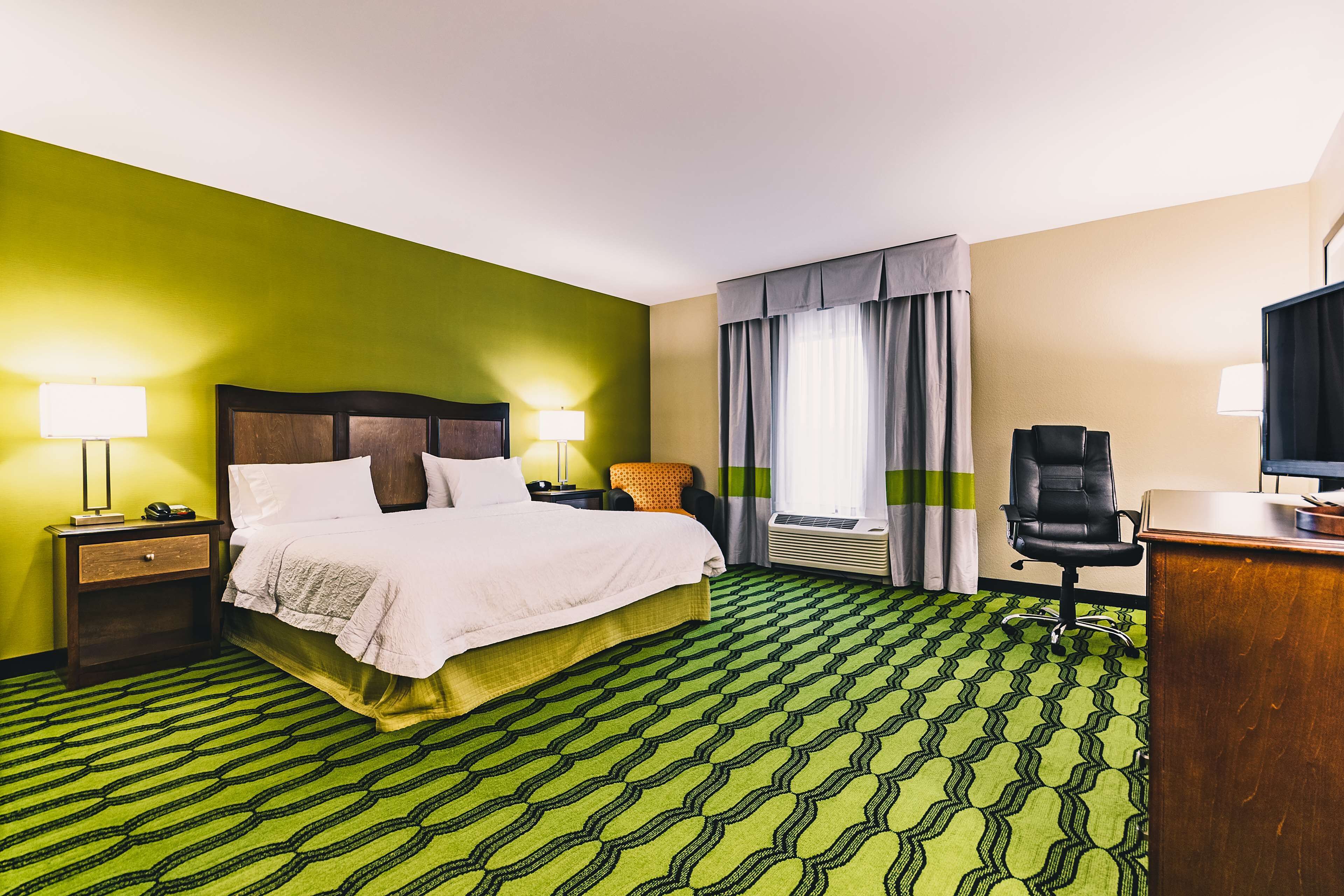 A hotel room with a bed, a desk, a chair, a lamp, and a window with curtains.