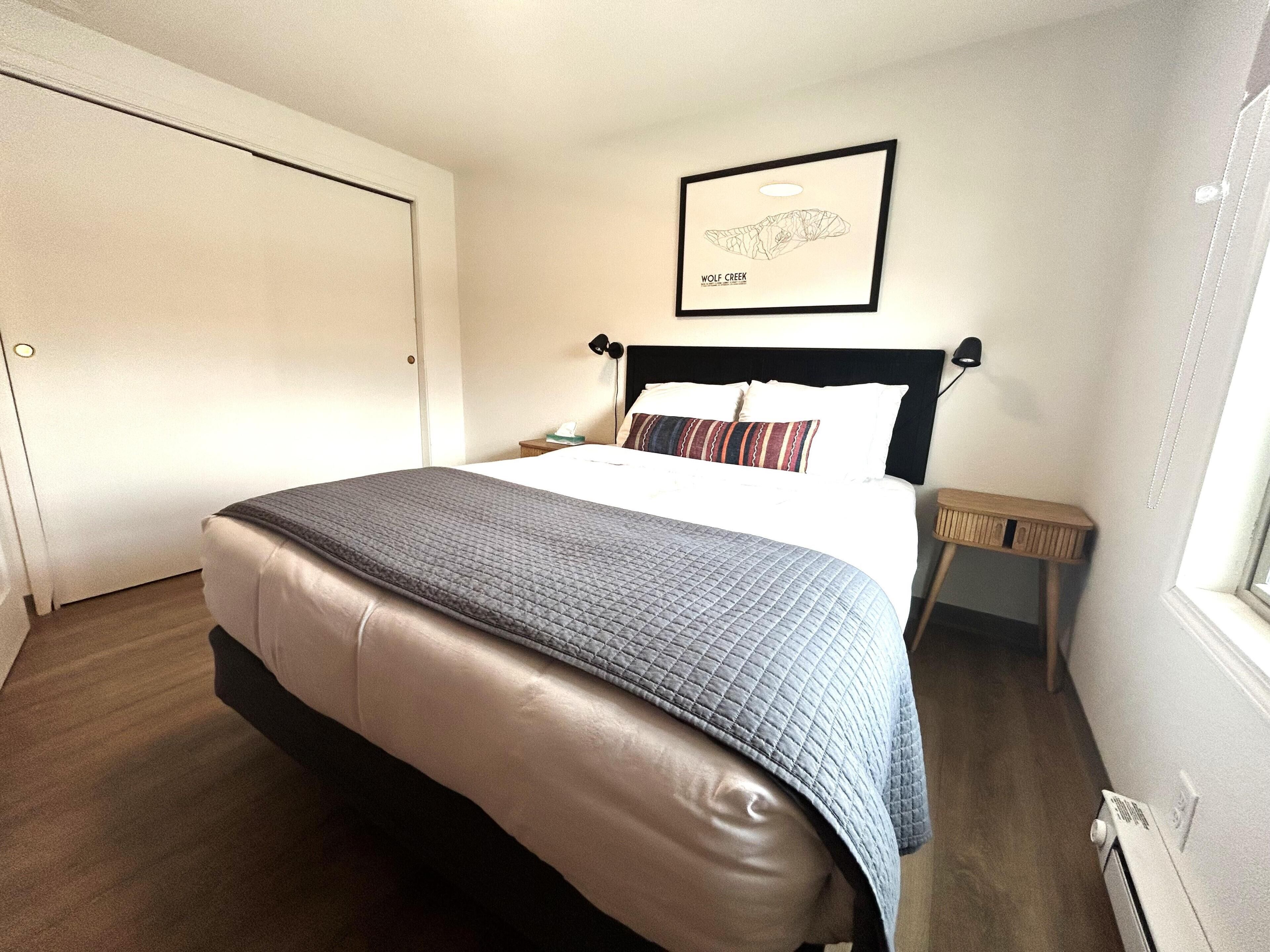 A neatly made bed with a quilt, a bedside table with a lamp, a framed picture on the wall, and a door to the left.