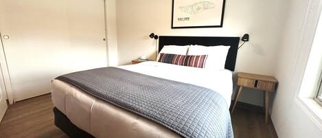 A neatly made bed with a quilt, a bedside table with a lamp, a framed picture on the wall, and a door to the left.