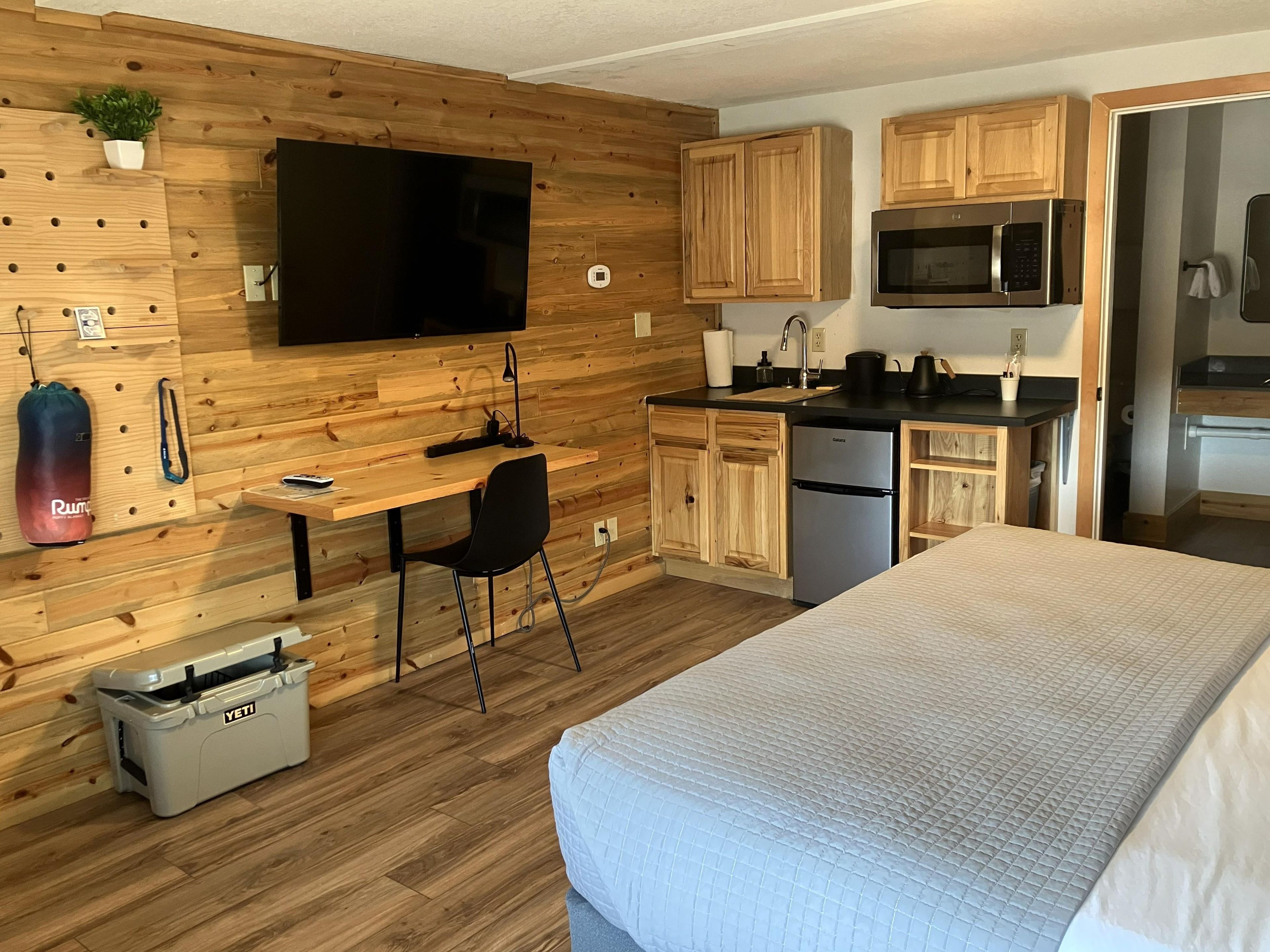 Habitación pequeña con cama, escritorio con silla, televisor montado en la pared, armarios de madera, microondas y refrigerador.