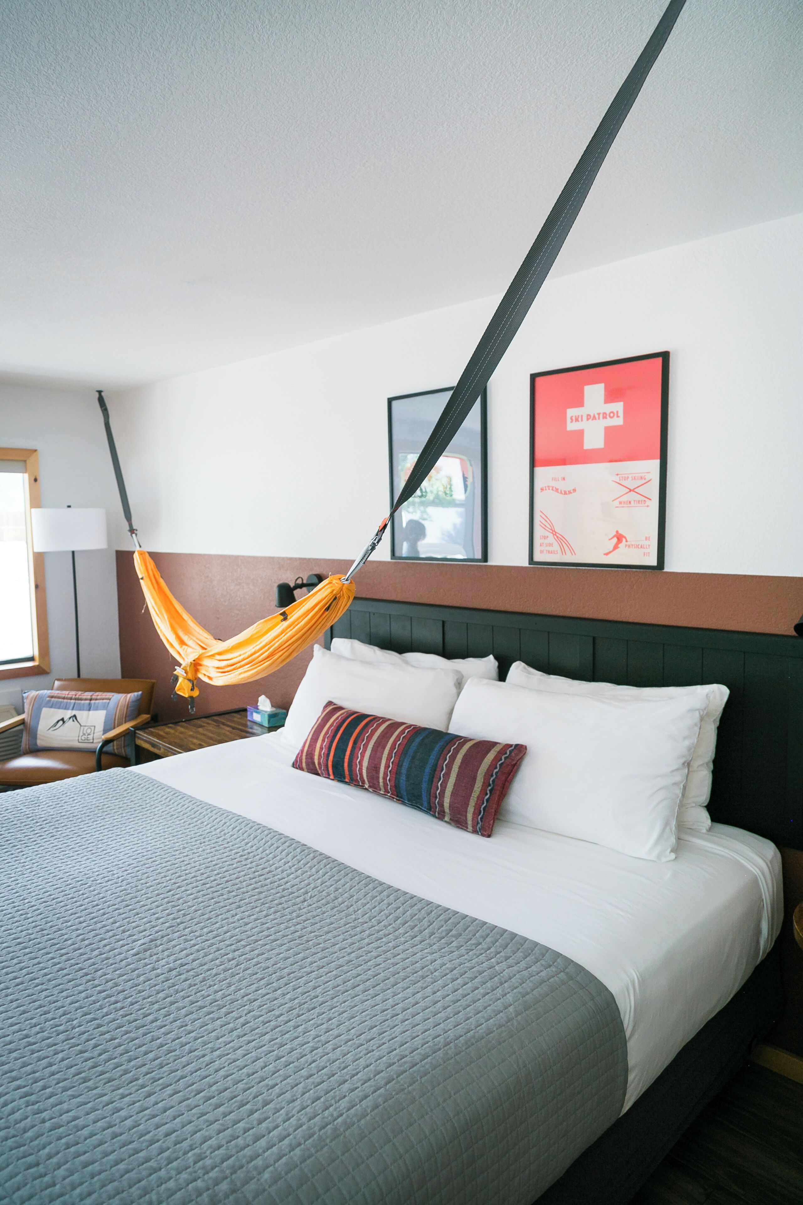 A hotel room with a bed, a hanging hammock, a nightstand, a lamp, and a poster on the wall.
