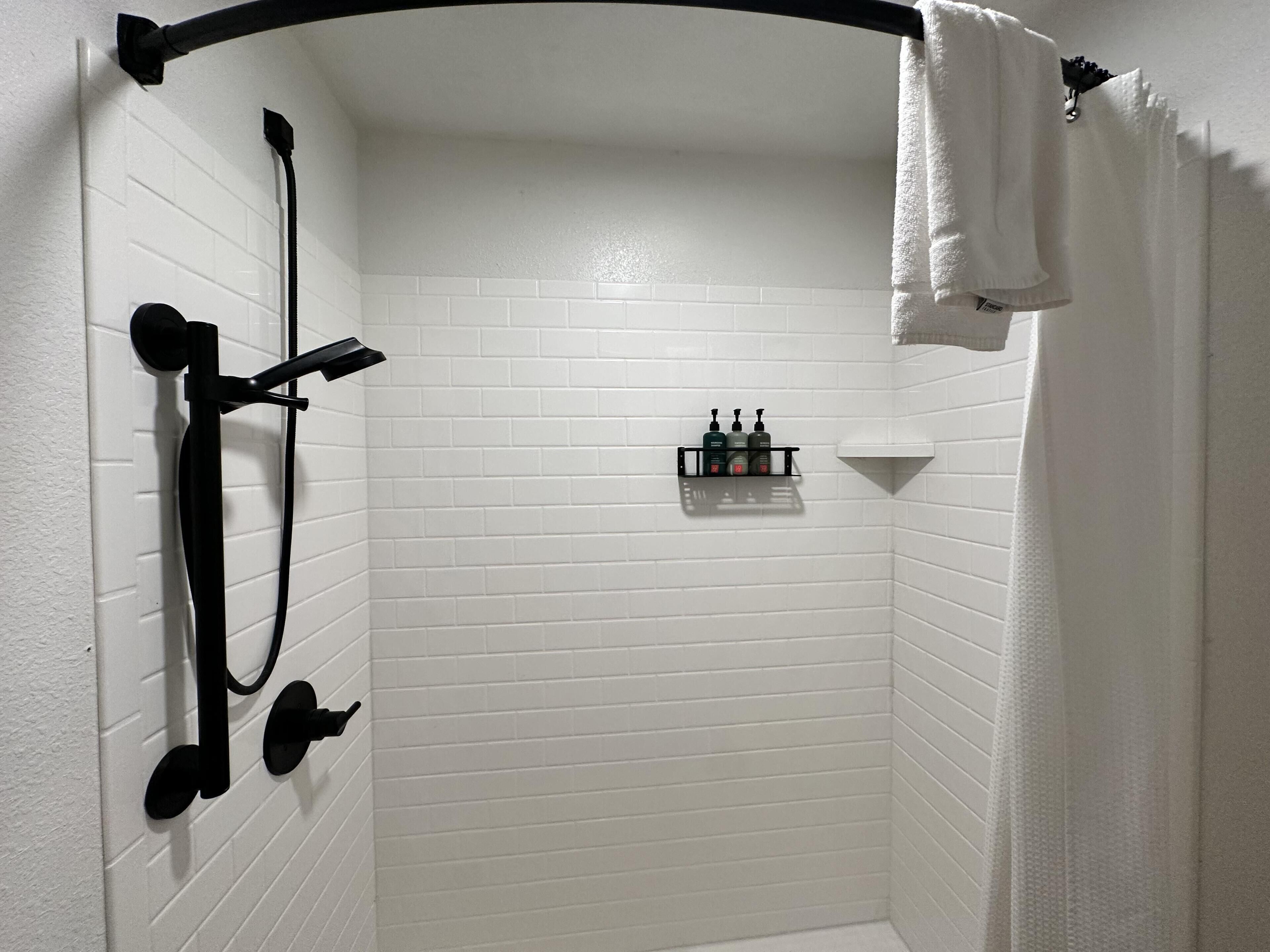 A corner shower with white tiled walls, a black showerhead, and a shelf with toiletries.