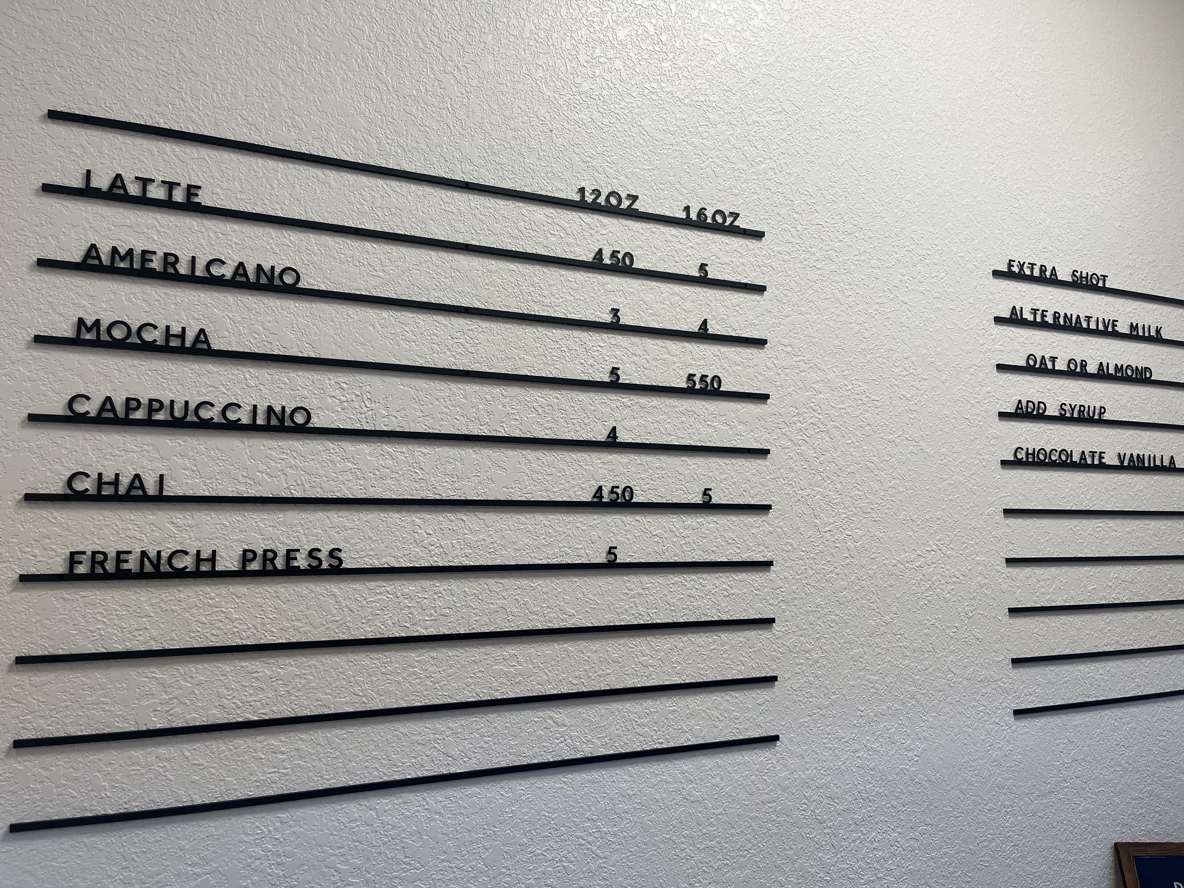 A wall with a menu listing coffee and drink options, including latte, americano, mocha, cappuccino, chai, and french press, with corresponding prices.